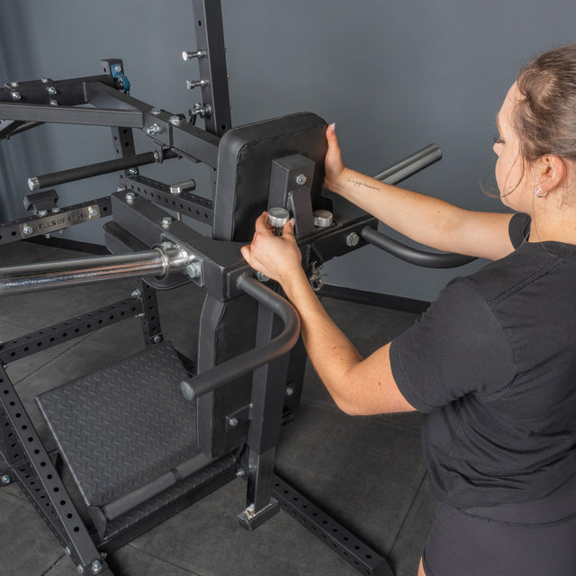A woman adjusts the settings on a Bells of Steel Pandemonium Squat – a large, black 3-in-1 pendulum squat, calf press, and Viking press machine with multiple handles and a padded backrest, in a gray-walled fitness room.