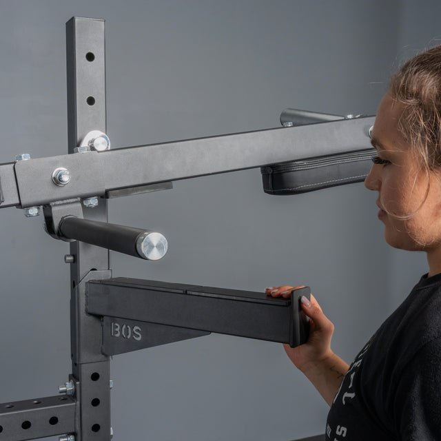 A person adjusts the black metal lever arm of the Bells of Steel Pandemonium Squat – 3-in-1 Pendulum Squat / Calf Press / Viking Press Machine in a home gym, gripping the padded handle. The background is plain gray.