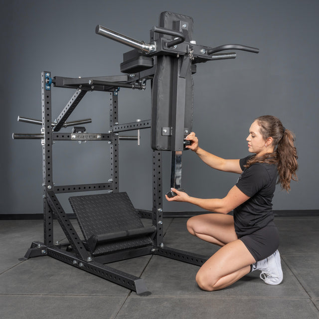 A woman in athletic wear adjusts the vertical pad of the Bells of Steel Pandemonium Squat – a 3-in-1 Pendulum Squat / Calf Press / Viking Press Machine, with sturdy metal supports and foot platforms, ideal for any home gym or intense workout.