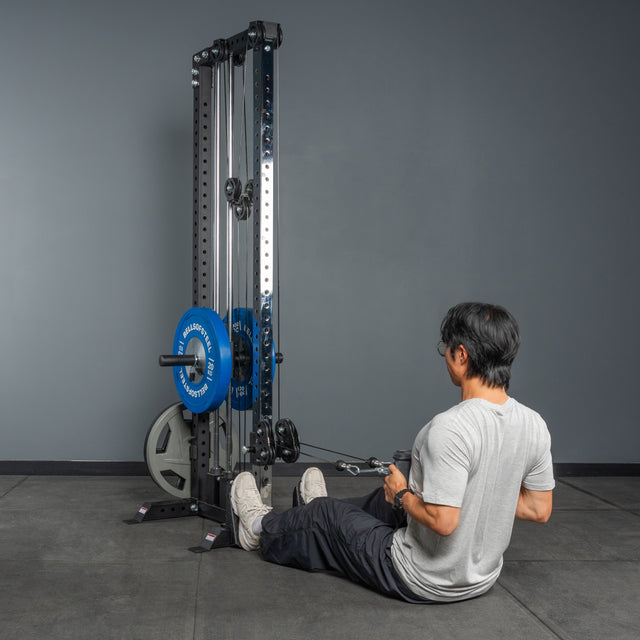 A person in a gray shirt and black pants uses the Bells of Steel Cable Tower with blue weight plates for cable machine exercises while sitting on the gym floor.