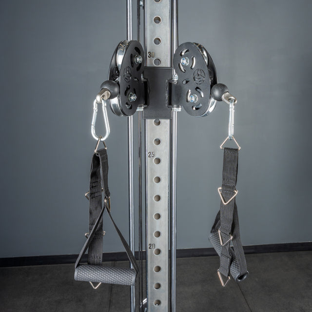 The Bells of Steel Cable Tower gym cable machine features adjustable pulleys, two black nylon handles with metal carabiners, and a visible weight stack—ideal for versatile exercises against a gray wall and floor.