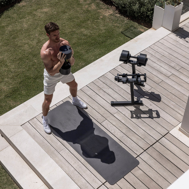 A man in white shorts and sneakers works out on a wooden deck, holding a dumbbell. Beside him, a black mat and the Nuobell NÜOBELL Adjustable Dumbbells rest neatly on the Nuobell NÜOBELL Floor Stand for space-efficient storage. Grass and plants line the deck.