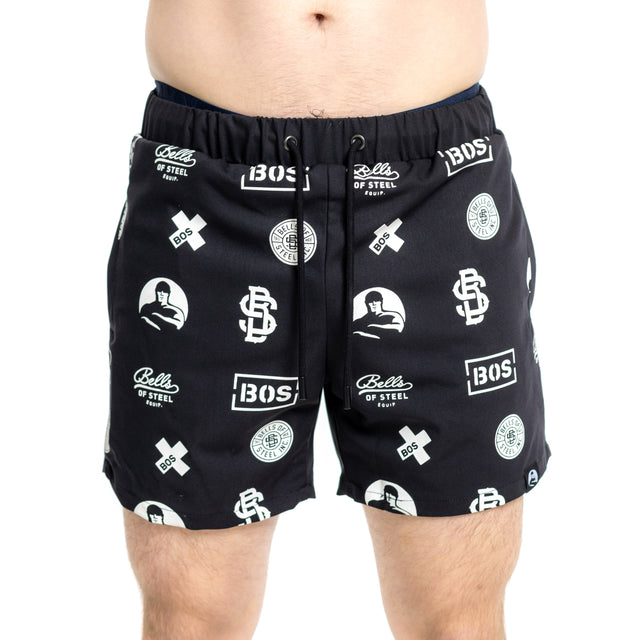 A person wears Bells of Steel Gym Shorts in black with white "Bells of Steel," "BOS," and logo prints. Made from stretchy fabric, these shorts have a drawstring waistband, sit above the knees, and show only the lower torso and upper legs.