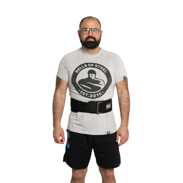 A bearded man with glasses, in a Bells of Steel T-shirt, black shorts, and a Bells of Steel Self Locking Belt for back support, stands facing forward against a white background—ready to start his fitness journey.