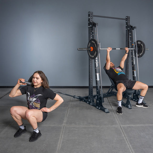 A woman squats with a barbell on her shoulder using the Bells of Steel Smith Machine - Hydra (3" x 3", ⅝" Holes), while a man bench presses on another Hydra rack behind her; both wear black workout clothes in a gym.