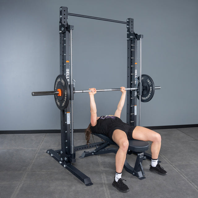 A person performs a bench press with weight plates on the Bells of Steel Smith Machine - Hydra (3" x 3", ⅝" Holes) in a gym, surrounded by a power rack and a nearby pull-up bar.