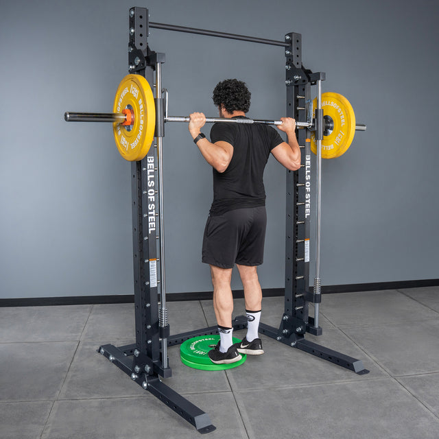 A person stands on a green weight plate inside a Bells of Steel Smith Machine - Hydra, getting ready to lift a barbell with yellow plates across their shoulders in a gym.