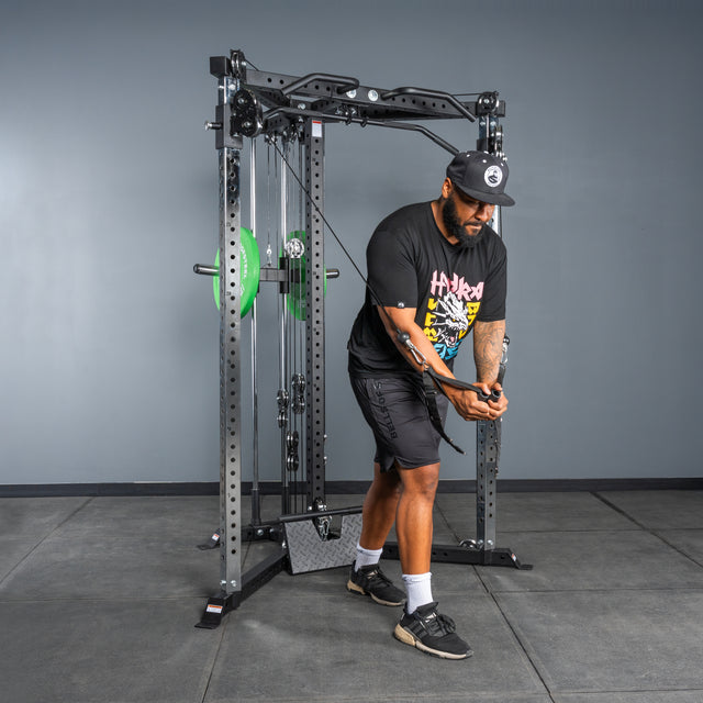 A man in athletic attire uses the Bells of Steel All-in-One Trainer for a cable crossover exercise in a gym, standing on rubber flooring against a neutral gray wall.
