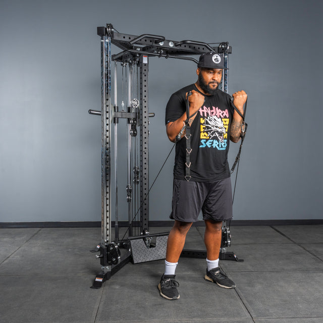 A man in athletic wear performs a biceps curl with resistance bands attached to the Bells of Steel All-in-One Trainer in a gym, standing on a gray floor against a plain gray wall.
