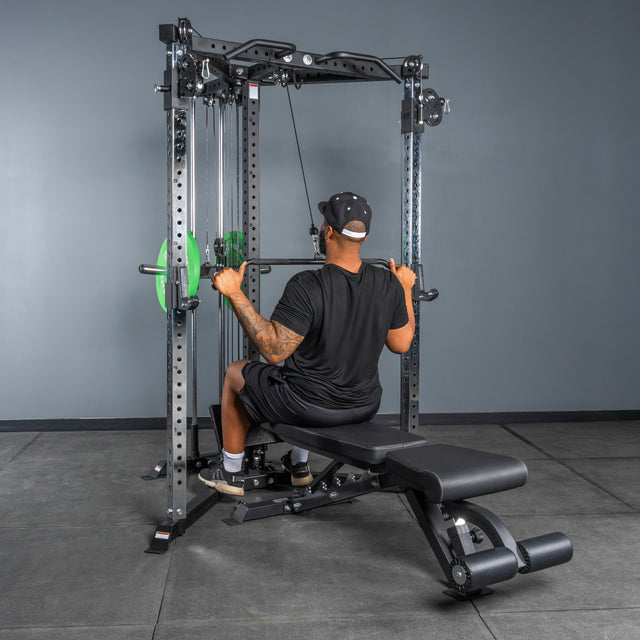 A person in black workout attire and cap uses the Bells of Steel All-in-One Trainer for a lat pulldown, seated on a bench and gripping the overhead bar in a gym.