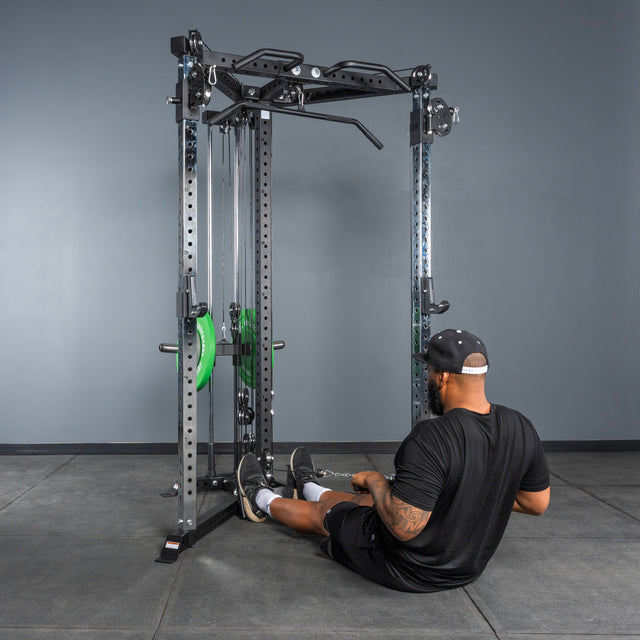 A man in black athletic wear sits on his home gym floor, facing the Bells of Steel All-in-One Trainer and gripping a handle as he prepares to perform a seated row exercise.