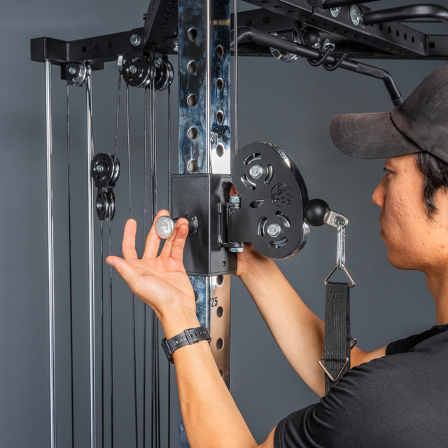 A person in a black shirt and cap adjusts the pulley on a Bells of Steel All-in-One Trainer, holding a pin near the adjustable mechanism of this versatile home gym machine.