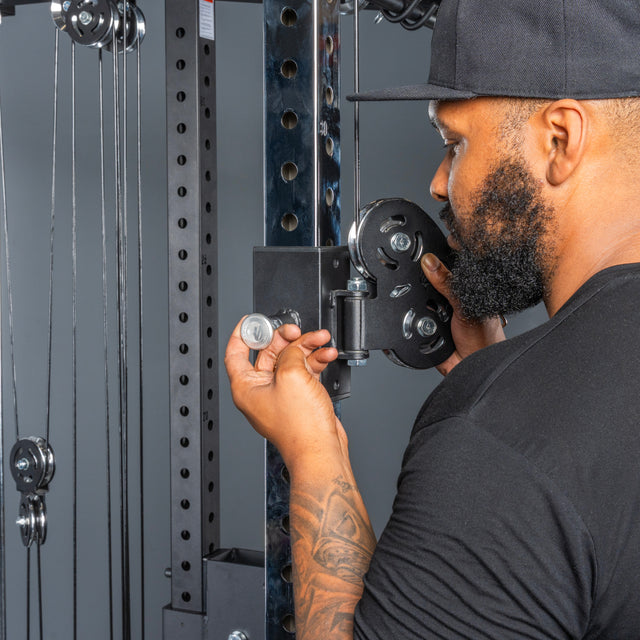 A bearded man in a black shirt and cap adjusts the settings on a Bells of Steel All-in-One Trainer at the gym, tightening a knob by hand.