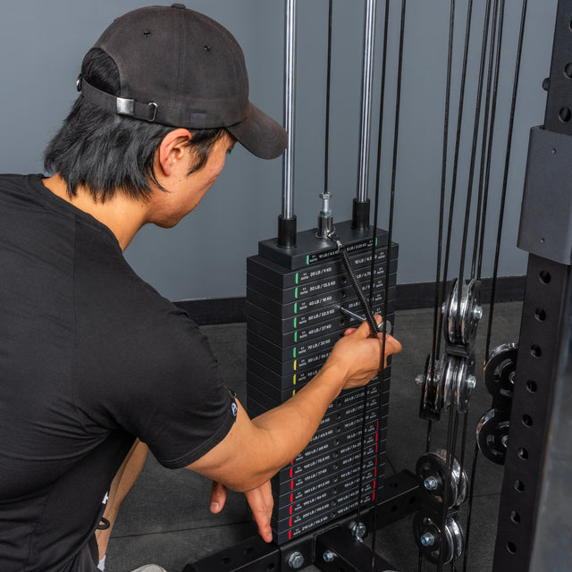A person in a black cap and shirt adjusts the weight stack pin on the Bells of Steel All-in-One Trainer in a gym.