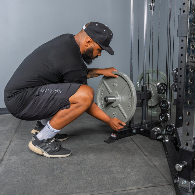 A man in athletic wear and a cap squats next to the Bells of Steel All-in-One Trainer, placing a weight plate onto the machine in a gym setting.