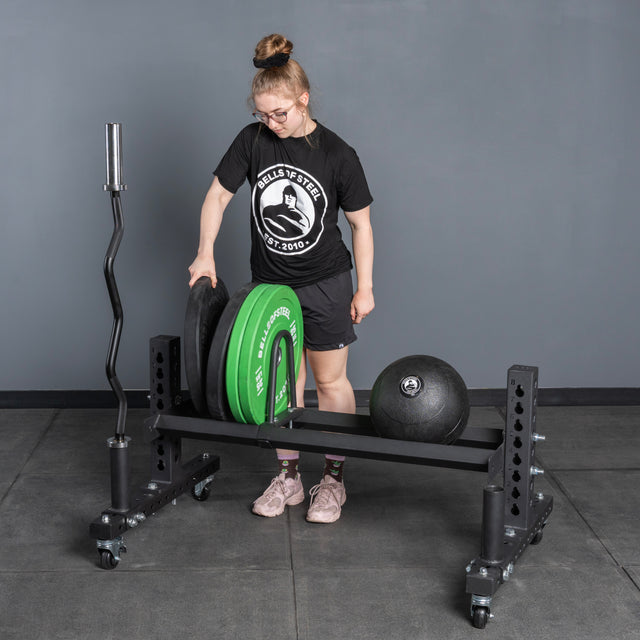 A person in athletic wear loads weight plates onto the Bells of Steel Rack Junctions - 3" x 3", with a barbell and weighted ball nearby on a gray gym floor—ideal for a versatile home gym setup against a plain gray wall.
