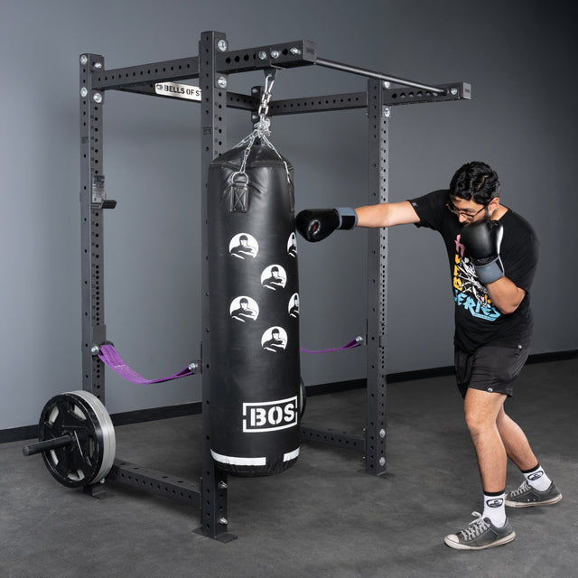 Wearing black boxing gloves, a person punches a black bag in a gym with gray walls, weights, and the Bells of Steel Rack Junctions - 3" x 3", an ideal customizable rack for any home gym setup.
