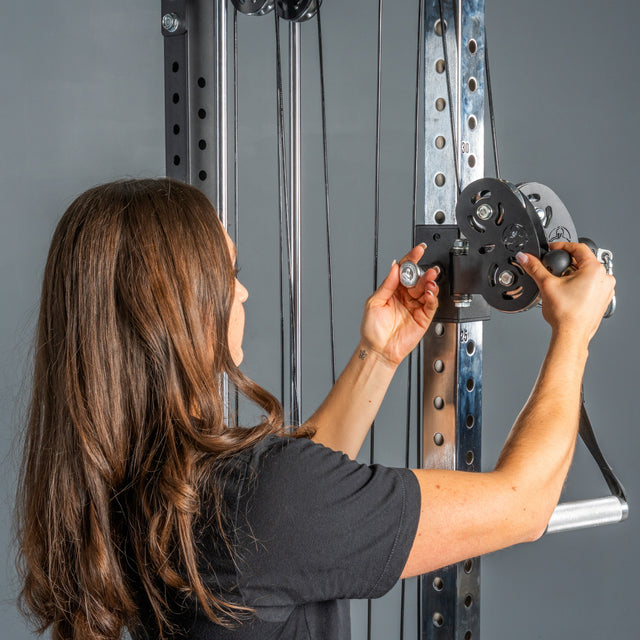A person with long brown hair adjusts a pulley system on the Bells of Steel Cable Tower, standing in front of the machine and attaching a black metal accessory near the weight stack with both hands.