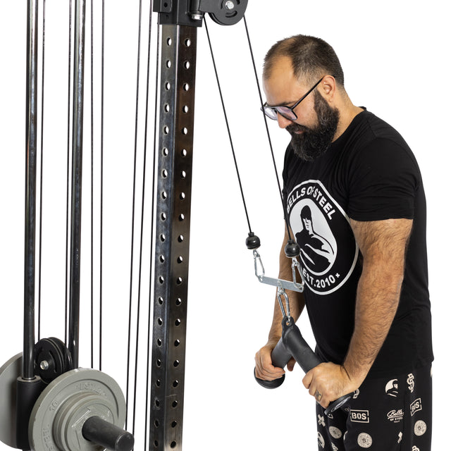 A man in a black t-shirt and glasses uses the Bells of Steel Fat Bar - Tricep Pressdown on a cable machine, gripping the handle with both hands and focusing intently on his workout to build grip strength.
