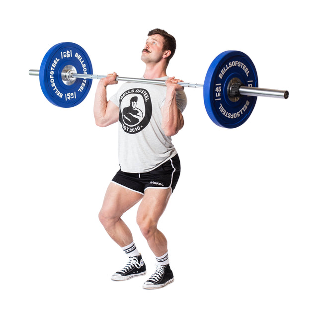 A man in a gray t-shirt and black shorts lifts a barbell with Bells of Steel Urethane Bumper Plates, preparing for an overhead Olympic lift. He wears sneakers and stands against a white background.