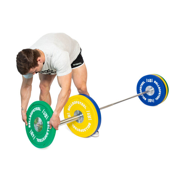 Wearing athletic clothing, a person bends over to load Bells of Steel Urethane Bumper Plates onto a barbell on the floor, preparing for weightlifting. The background is completely white.