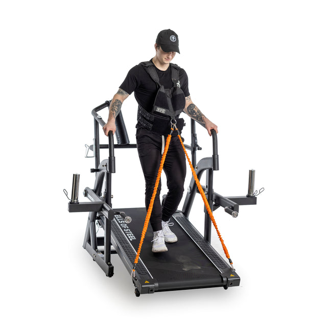 A person in a black Bells of Steel weighted vest walks on a treadmill with orange resistance bands. They are dressed in black and white, showcasing tattoos on their arms as they grip the treadmill handles, highlighting an adjustable workout setup.