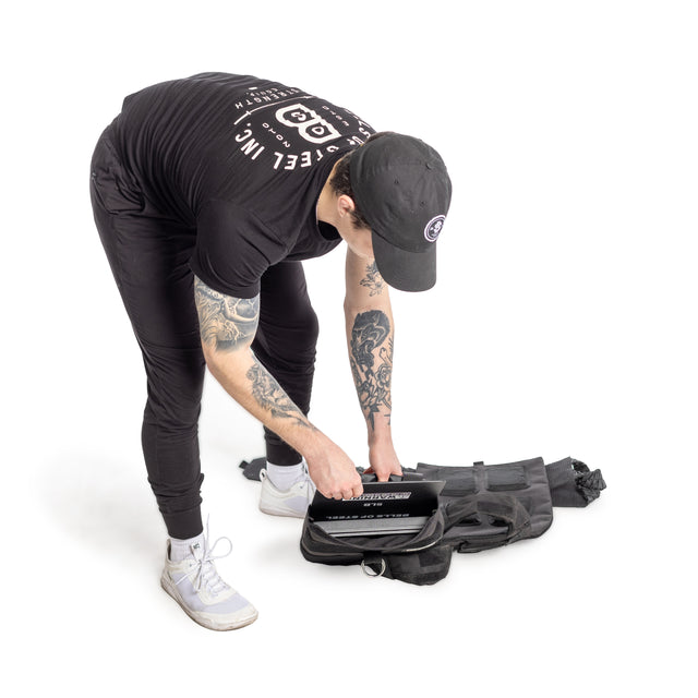 A person in black athletic wear and a cap, displaying tattooed arms, bends over to place a laptop into an open black backpack on the floor. Next to the bag is a Bells of Steel Weighted Vest with training plates, emphasized by the white background.