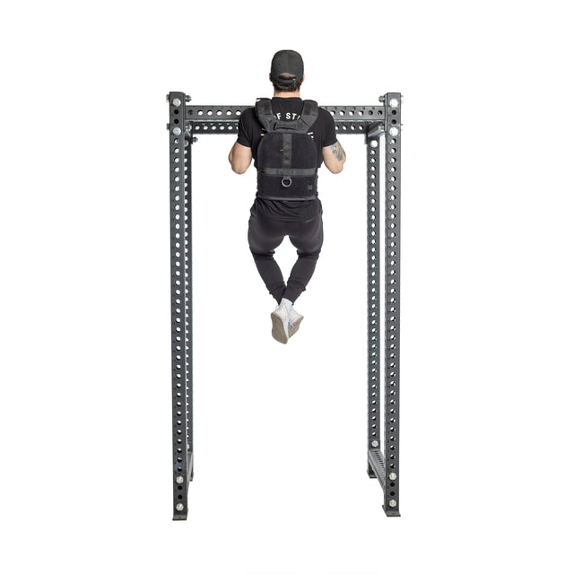 A man in a black cap and workout clothes performs a pull-up using a Bells of Steel Weighted Vest on a metal bar, showcasing his strength from behind with legs slightly bent, enhanced by the vest's adjustable training plates.