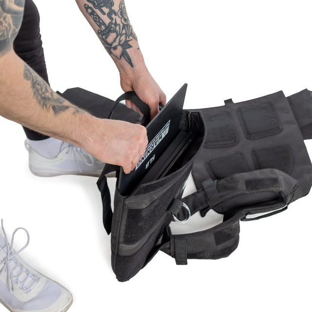 A person with tattooed arms inserts a laptop into an adjustable black Bells of Steel Weighted Vest laid on a white surface. They wear dark pants and white sneakers, using the vest's weight training plates for versatile fitness.