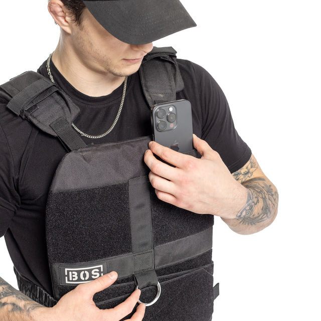 A tattooed individual wearing a black Bells of Steel Weighted Vest, designed for training plates and featuring a logo and metal ring on the front, adjusts the pocket while holding a smartphone. They complete their look with a chain necklace and cap.
