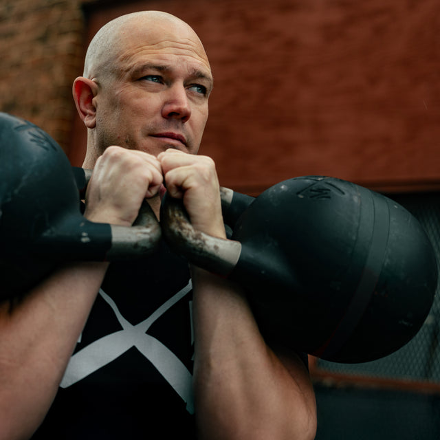 A bald man with a serious expression holds the Bells of Steel 48KG Extension Kit for Adjustable Kettlebell – Wildman Poison Pill at chest height outdoors, wearing a black shirt with a white X design. A red brick wall is in the background.