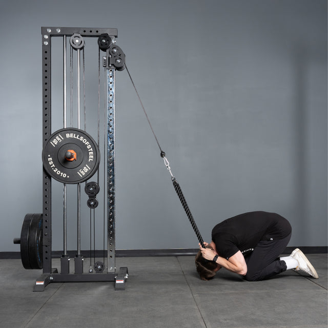 Wearing black workout gear, a person kneels in child's pose, using the Bells of Steel Extra Long Tricep Rope Extension (137cm) attached to a weight machine to perform a cable crunch exercise.