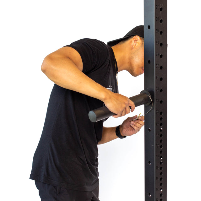 A person in a black shirt and cap uses a wrench to tighten a bolt on the Bells of Steel Utility Horn Rack Attachment, preparing it for Olympic weight plates. The plain white background emphasizes their precise adjustments.