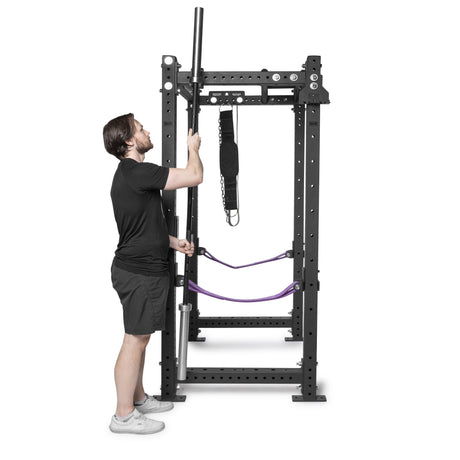A man in a black shirt and shorts adjusts a barbell on a black power rack equipped with Bells of Steel Hanging Barbell Holders, safety straps, and dip belt, set against a white background.