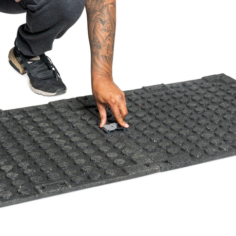 A man installs a Bells of Steel Rubber Flooring Gym Mat 24" x 24" by placing a small connector piece between tiles. Only their hand, arm, shoe, and part of the black textured mat appear against a white background.