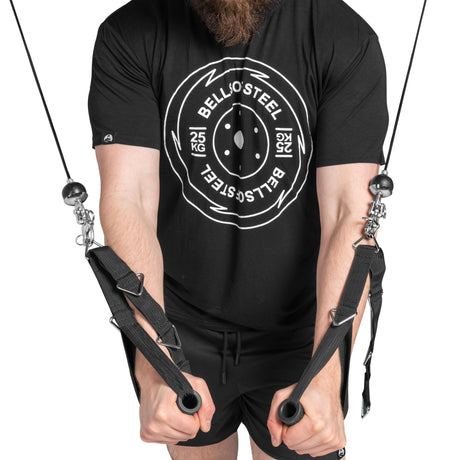 A person in Bells of Steel black T-shirt and shorts is exercising on a cable machine equipped with Swivel Shackles, gripping black handles and pulling them downward, highlighting their torso and arms while demonstrating their gym gear.