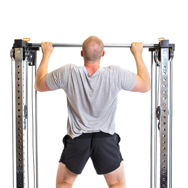 Sporting a gray T-shirt and black shorts, a person with a shaved head performs pull-ups on the Bells of Steel Cable Tower Squat Stands in front of a white background, emphasizing the gym equipment.