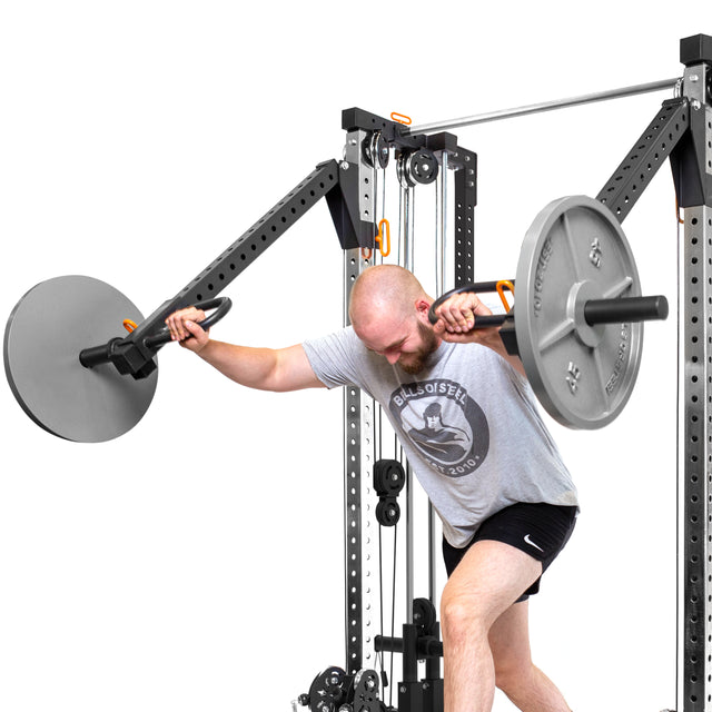 A man in a gray shirt and black shorts exercises with Bells of Steel Cable Tower Squat Stands, leaning forward and extending his arms. The plain white background provides an ideal minimalist setting for a versatile home gym routine.