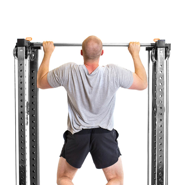 Someone in a gray T-shirt and black shorts is performing a pull-up on the Bells of Steel Cable Tower Squat Stands, echoing versatile home gym equipment. The view from behind against a stark white background emphasizes their strength and focus.