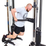 A man uses Bells of Steel's Cable Tower Squat Stands, doing a dip in gray T-shirt, black shorts, and sneakers with colorful socks. The metallic equipment showcases pulleys and weight stacks against a white backdrop.