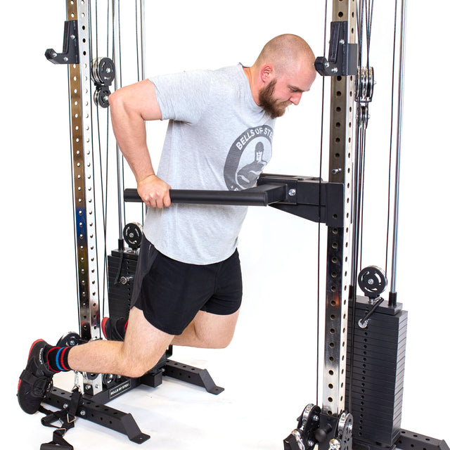 A person in a gray T-shirt and black shorts uses the Bells of Steel Cable Tower Squat Stands for dips, possibly in their home gym. They're positioned between two weight stacks with feet on a raised platform. The background is white.