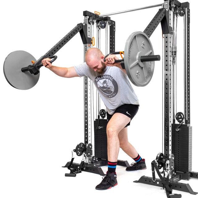 A man workouts on the Bells of Steel Cable Tower Squat Stands, with a barbell weight on an arm attachment. He's in a gray t-shirt, black shorts, and sneakers. The machine features weight stacks and adjustable arms, ideal for any home gym against a plain white background.