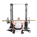 A man, showcasing his dedication to his home gym routine, performs a squat with a barbell using Bells of Steel's Cable Tower Squat Stands. He's dressed in a gray shirt, black shorts, and colorful socks.