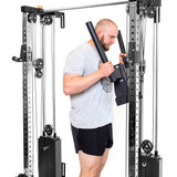 A person is setting up a Bells of Steel Cable Tower Squat Stands in their home gym. In gray t-shirt and black shorts, they hold bars, focused on their workout. The machine boasts adjustable weights and pulleys against a white floor backdrop.