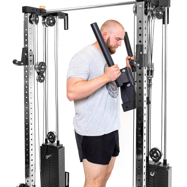 A person is setting up a Bells of Steel Cable Tower Squat Stands in their home gym. In gray t-shirt and black shorts, they hold bars, focused on their workout. The machine boasts adjustable weights and pulleys against a white floor backdrop.