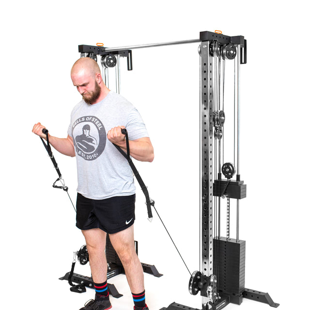 A bearded person exercises using Bells of Steel's Cable Tower Squat Stands. They're pulling handles attached to the cables, dressed in a gray T-shirt, black shorts, and striped socks against a plain white background.