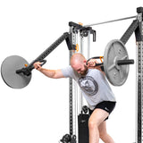 A bearded person is lifting a barbell with both hands in a gym, wearing a gray T-shirt and black shorts. The Cable Tower Squat Stands by Bells of Steel are black and silver against the plain white background.