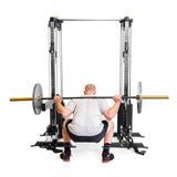 An individual in a gray shirt and black shorts performs a squat with a barbell from Bells of Steel's Cable Tower Squat Stands, set in front of plain white walls suggesting a minimalist home gym.