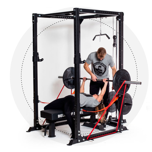 A man helps a woman bench press with resistance bands on the Home Gym Builder. Both wear logoed gray T-shirts. The setup features a padded bench and barbell with weight plates, crafted by related_to_7423949242420.