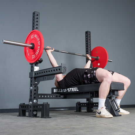 A person lies on a Bells of Steel Manticore Two Post Converter Bench, lifting a barbell with red plates in a gym. They wear a black shirt, black shorts, white socks, and beige sneakers.
